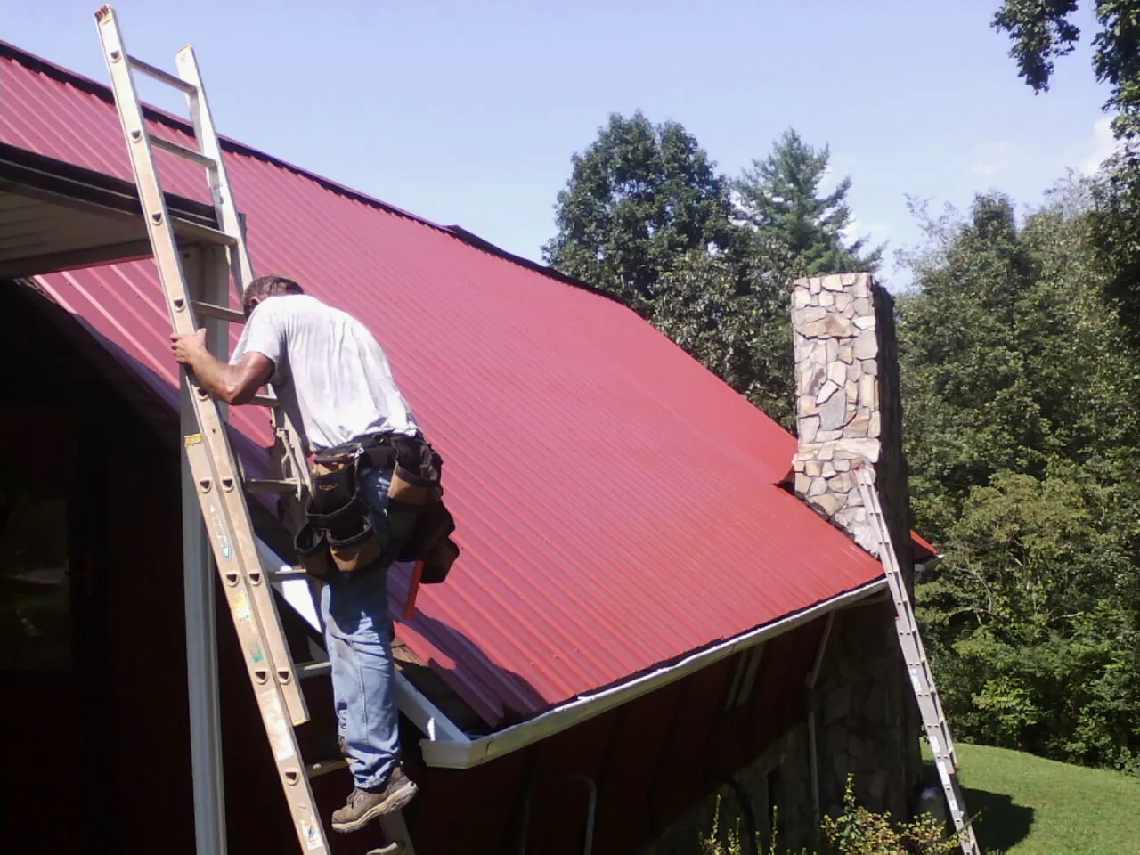 Roofer on a ladder beside a red metal roof and stone chimney — Soffit & Fascia Repair by Roofing Brothers in South Dayton